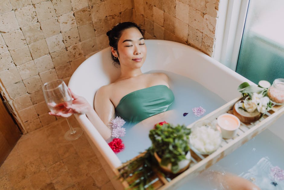 Asian woman enjoys a peaceful flower bath, holding a glass of wine, surrounded by candles for a serene spa experience.